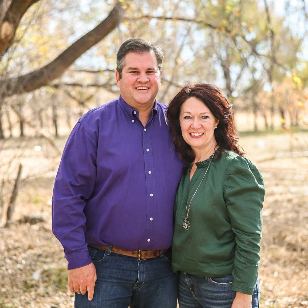 A smiling man and woman stand close together outdoors in a sunlit, wooded area with dry grass and trees in the background. The man wears a purple shirt; the woman wears a green top.