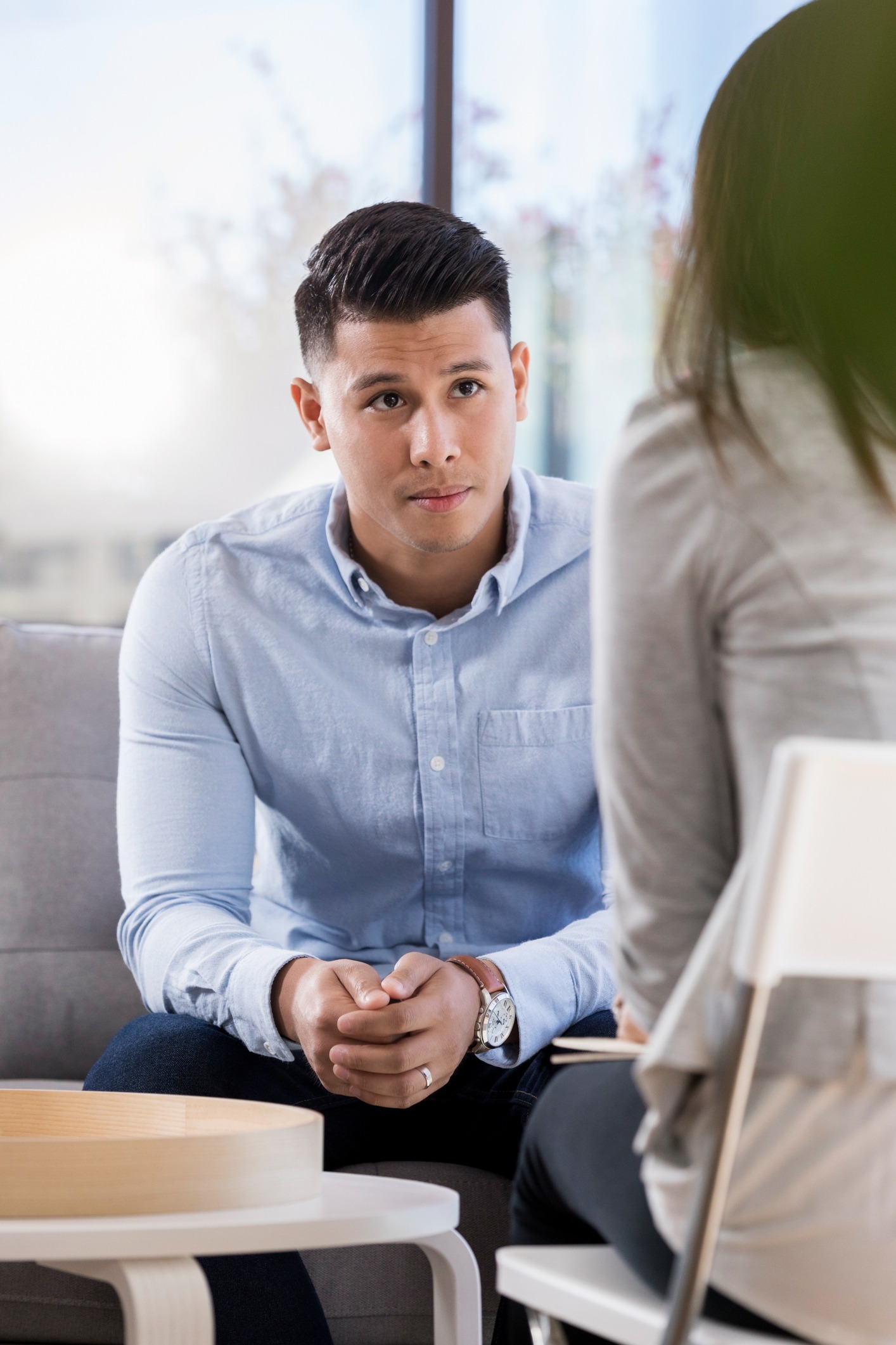 A man in a light blue shirt sits on a couch, listening attentively to a woman seated across from him. They appear to be engaged in a serious conversation in a bright, modern room.