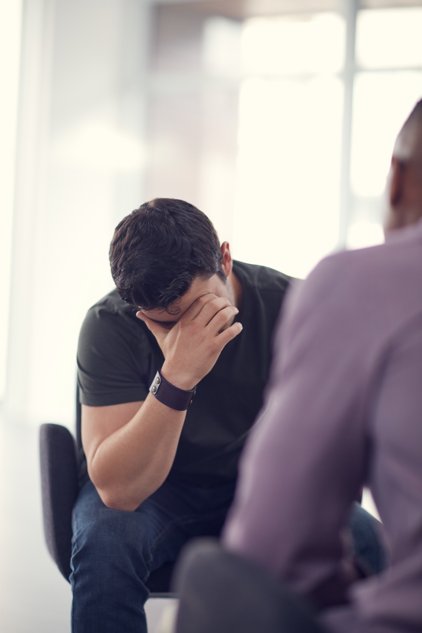 A man sits with his head bowed and hand covering his face, appearing distressed, while another person, blurred in the foreground, listens during a conversation in a bright room.