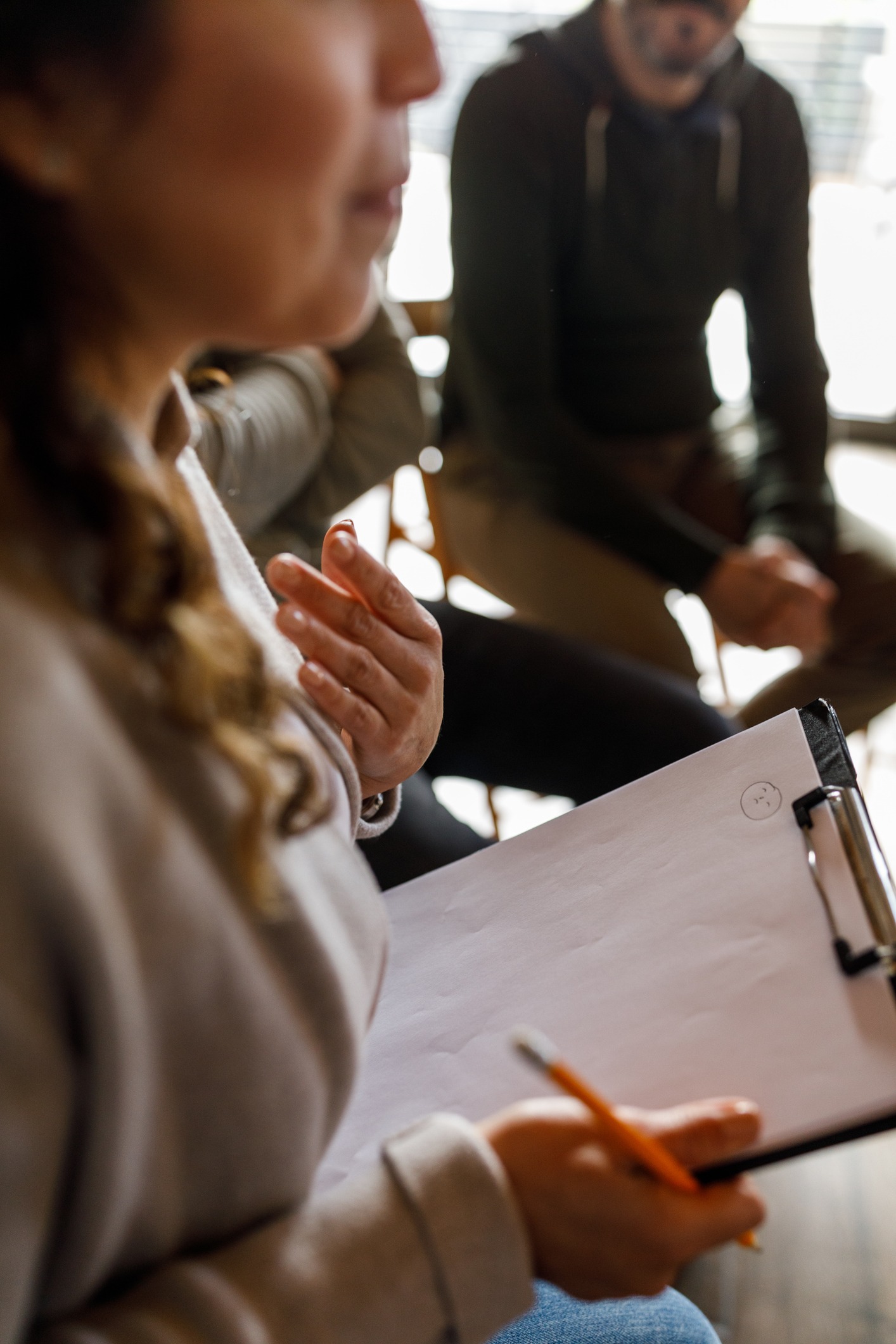 A person holding a pencil and clipboard with blank paper sits among others in a casual group setting, appearing to take notes or prepare to write. The focus is on the clipboard and the person's hand.