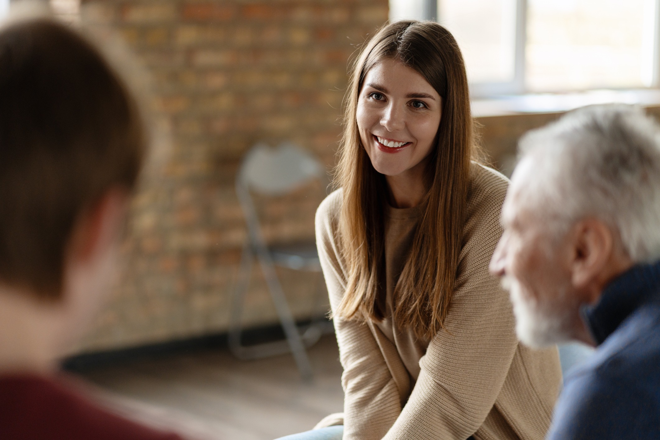 A young woman with long brown hair smiles while sitting and talking with two people in a bright room with exposed brick walls.